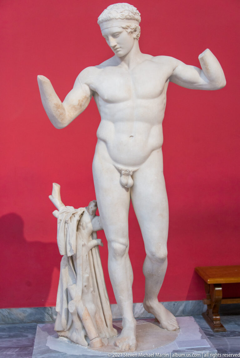 Large marble nude male statue with raised broken arms and a draped support beside one leg.