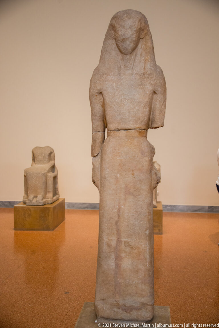 Large headless stone male figure with missing limbs displayed on a museum plinth.