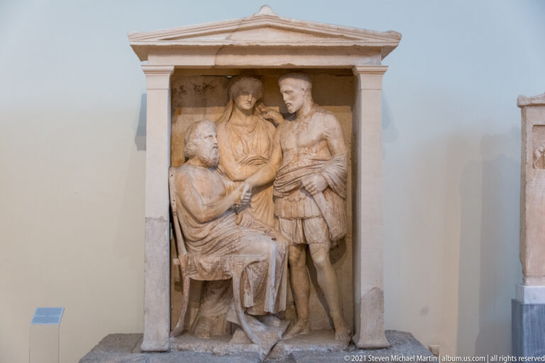 Marble grave stele with a seated figure, a standing armored man, and a third figure beneath a pediment.