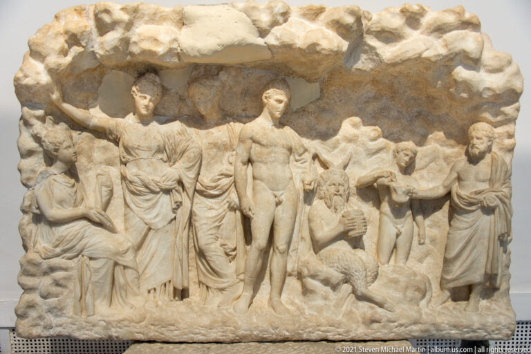 Weathered marble relief with a central nude man, several draped figures, smaller figures, and animals.