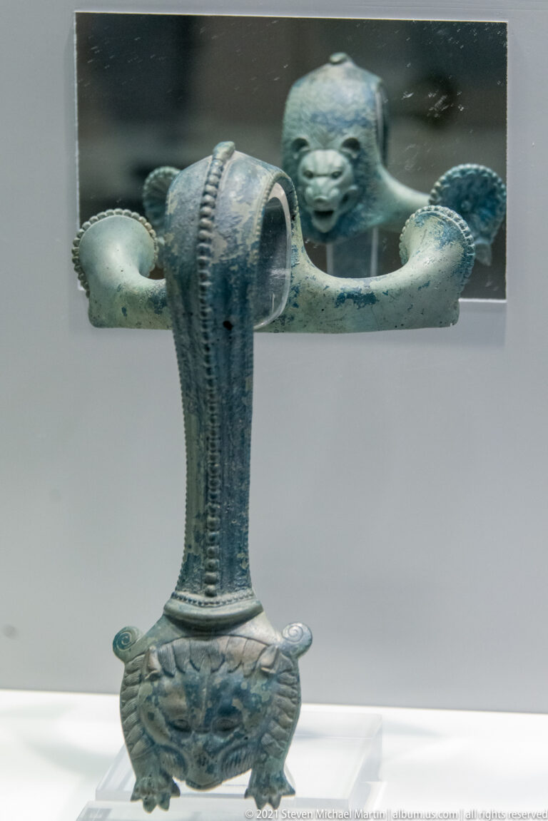 Tall bronze attachment with a ribbed stem, mask-like lower terminal, and broad transverse top.