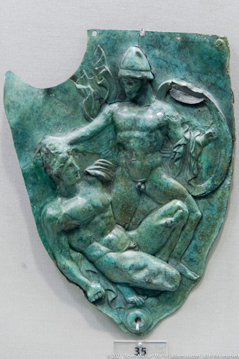 Irregular green bronze relief fragment with an armed male figure, animals, and shield-like forms.