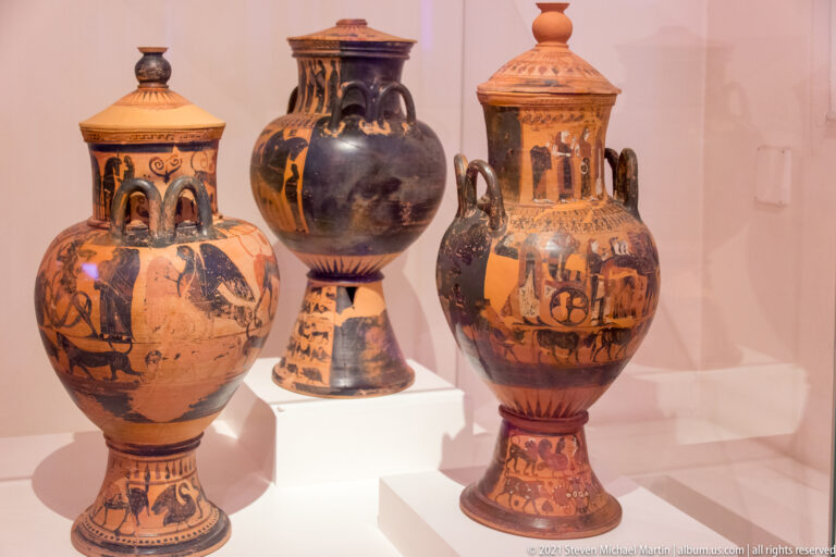 Three large ancient Greek painted ceramic vessels with tall necks, broad bodies, and geometric and figural decoration.