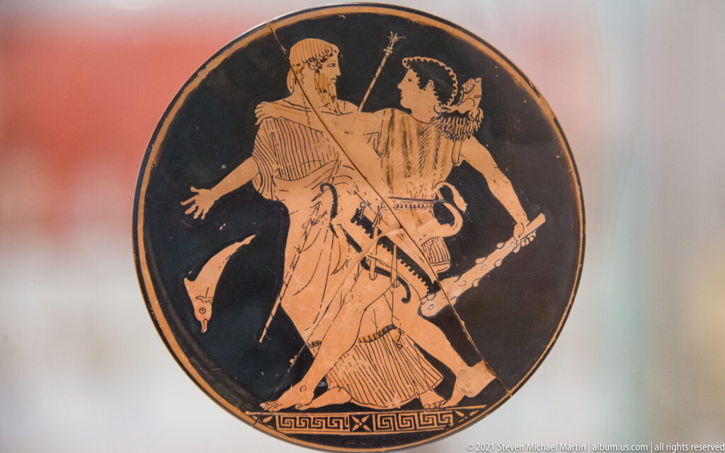Circular red-figure tondo with two standing male figures, one extending an arm toward the other, above a meander border.