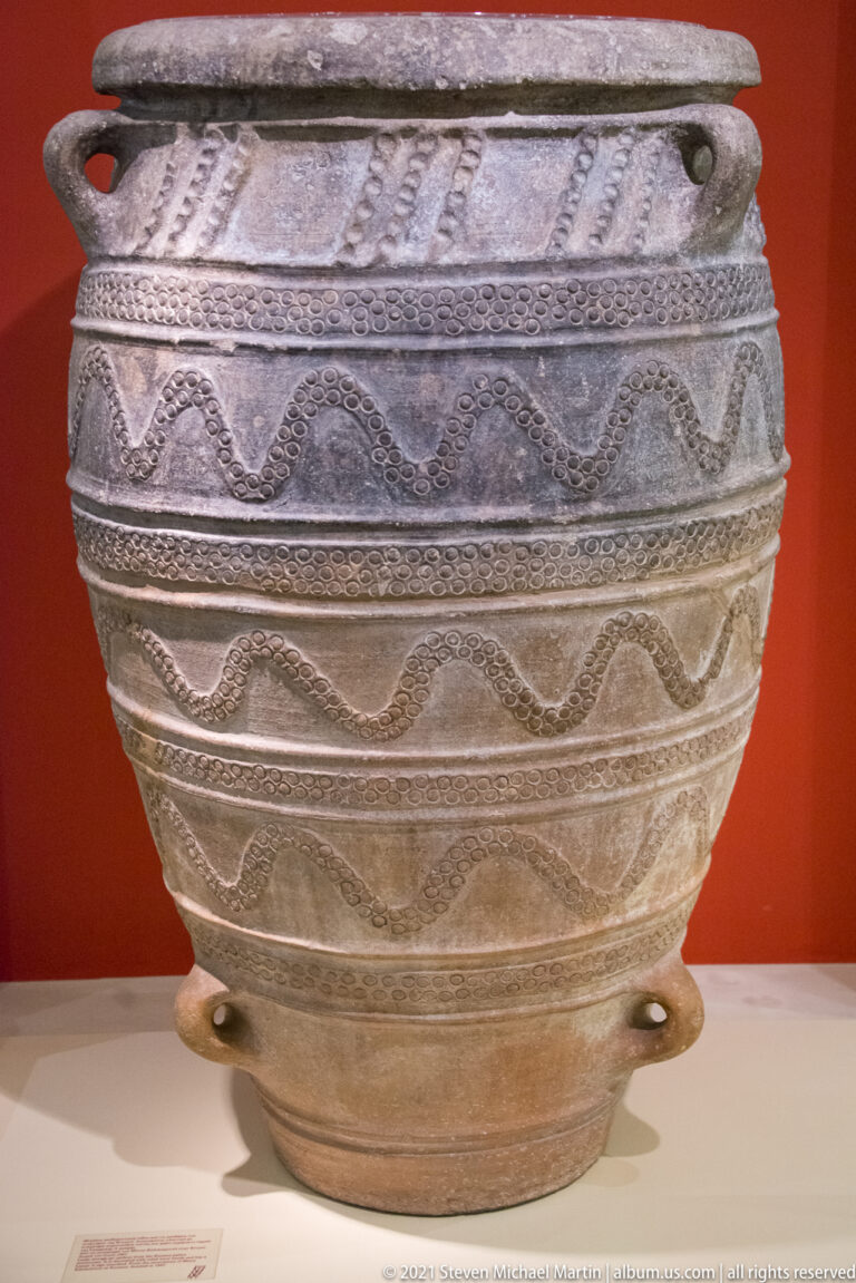 Large rounded ceramic vessel with repeated incised wavy bands, textured borders, and small projections near the base.