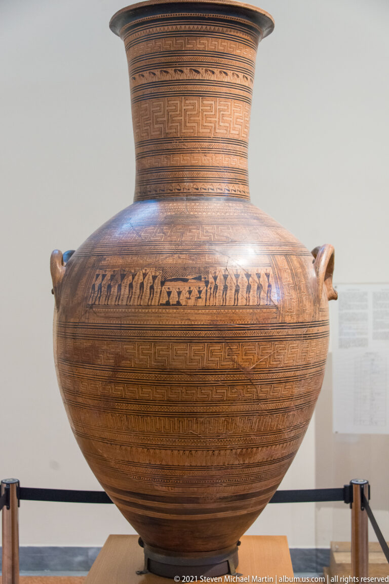 Large Greek ceramic vessel with geometric bands, figured frieze, tall neck, and two side handles.
