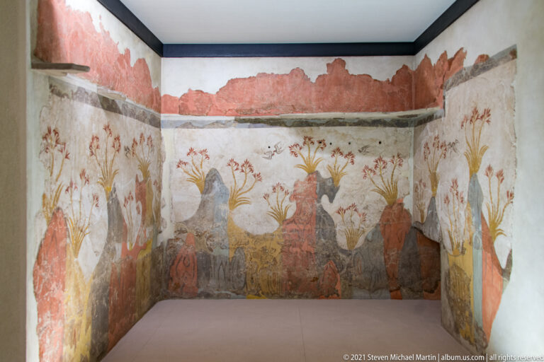 Reconstructed room displaying the Spring Fresco from Akrotiri, with red rocky forms, flowering lilies, and small birds across adjoining walls.