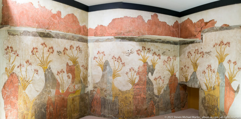 Alternate corner view of the Akrotiri Spring Fresco with lilies, red rocky forms, and small birds continuing across two walls.