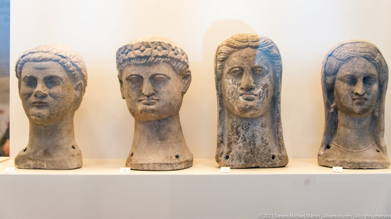 Four ancient sculpted heads displayed side by side on a museum shelf, with varied hairstyles and facial types.