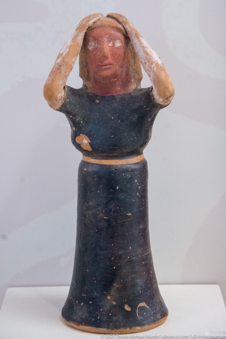 Small painted female figurine with both hands raised to her head, red face and arms, and a dark bell-shaped garment.
