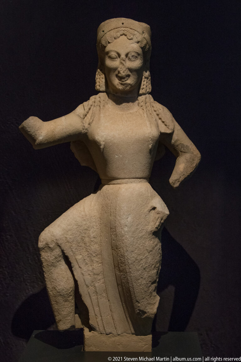 Archaic-style female statue with one arm extended, the other at the waist, and a pleated draped skirt.