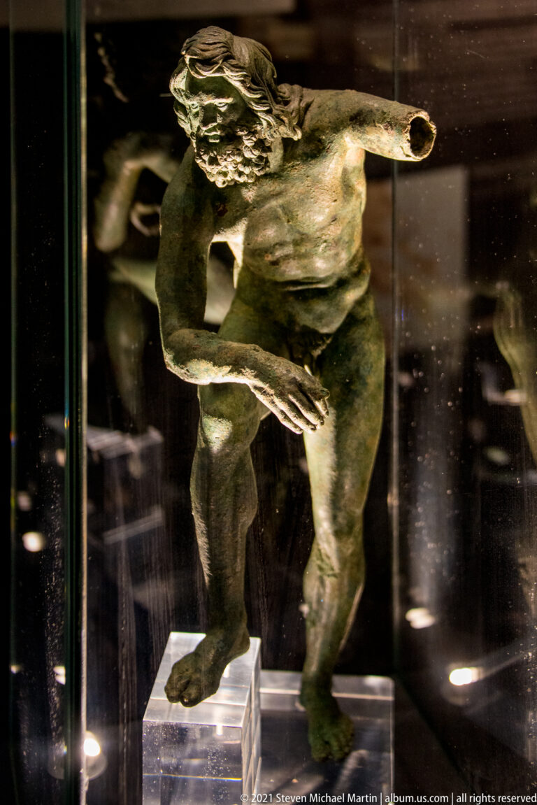 Bronze satyr- or Silenus-like male figure bending forward with shaggy hair and one arm extended.