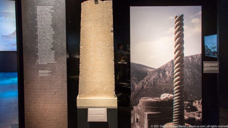 Museum display with a tall inscribed stone monument beside a twisted column-like reconstruction and interpretive panels.
