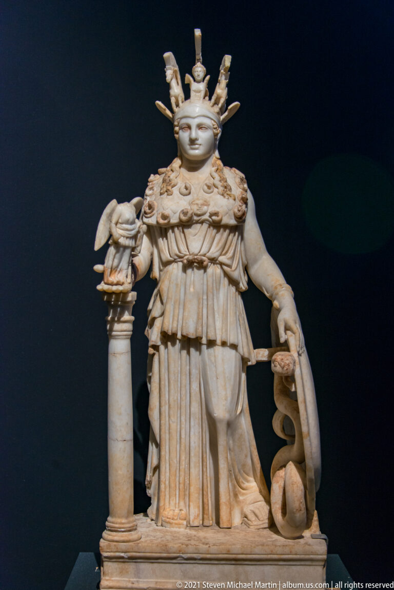 Helmeted Athena statuette in a long garment, holding a shield beside a small figure on a column.