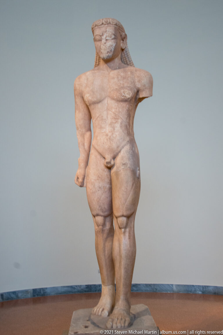 Front-facing nude male kouros with long hair, advanced leg, and partly missing arms.