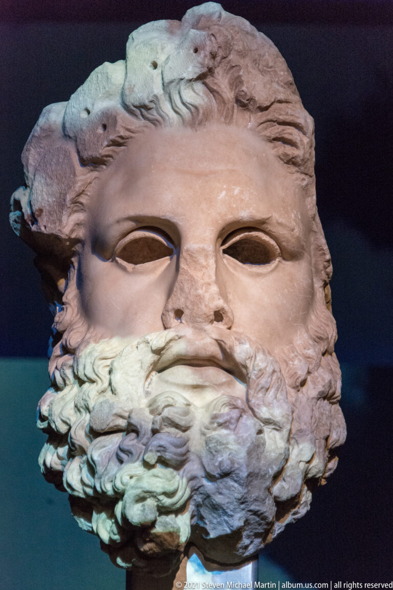 Colossal bearded male face with deep-set eyes, wavy hair, and a full curling beard in dramatic museum lighting.