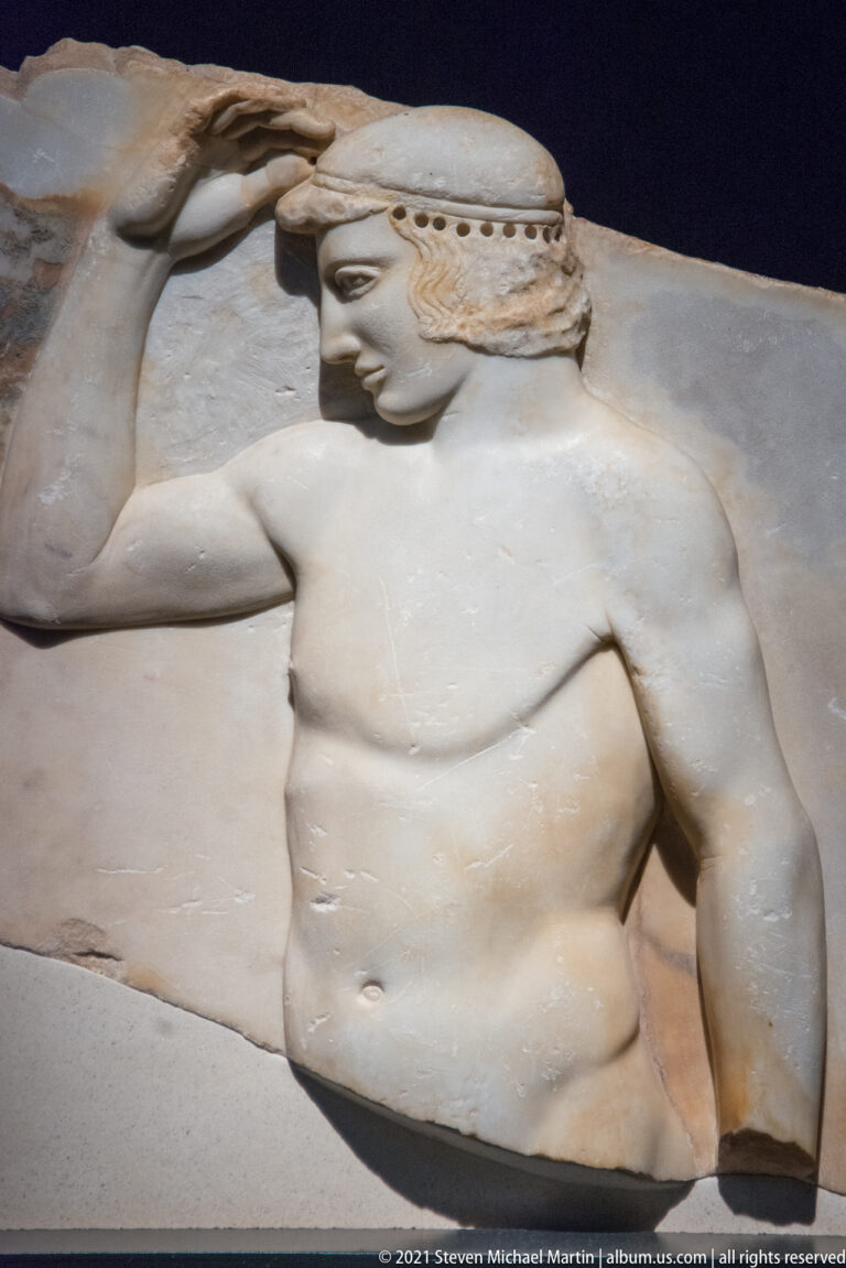 Fragmentary marble relief of a muscular male figure wearing a headband, with one arm raised behind him.