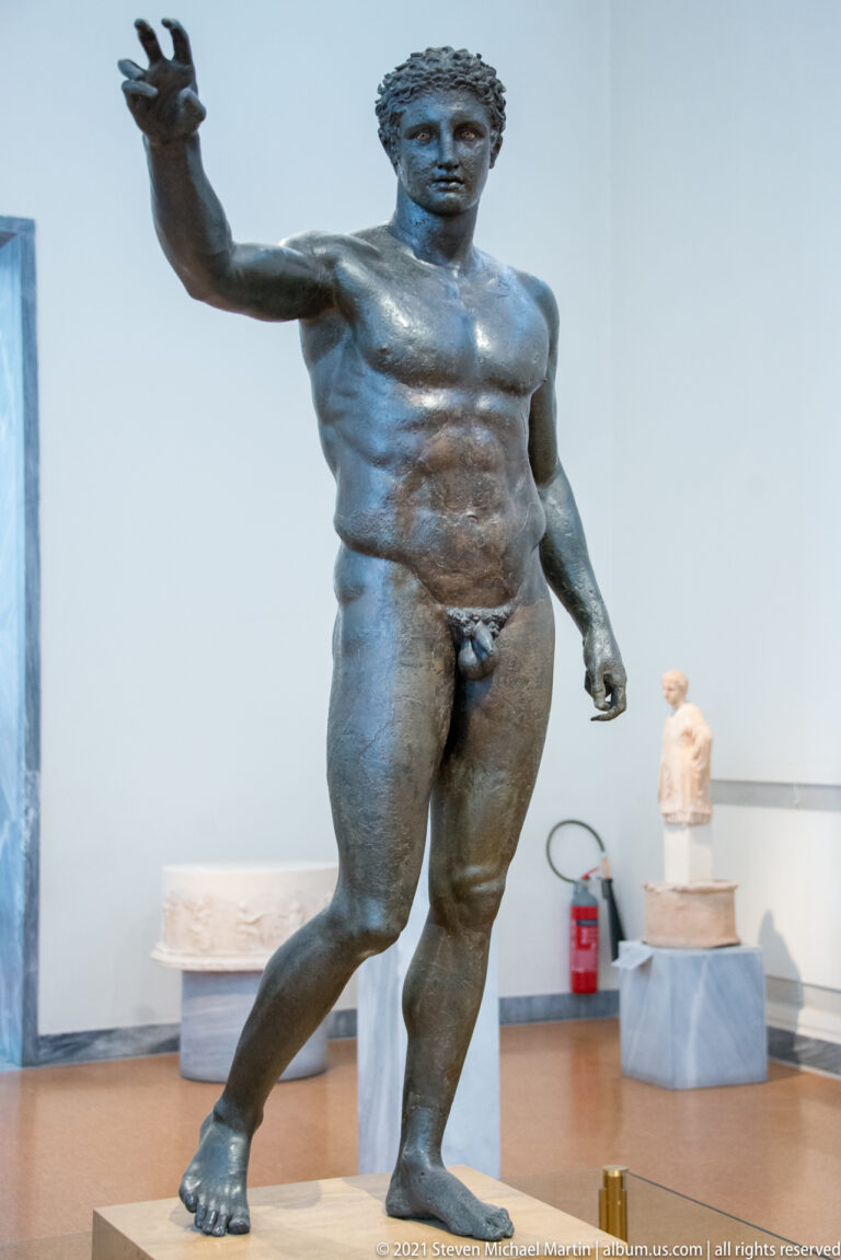 Life-size bronze nude male statue with short hair, one arm extended outward, and weight shifted onto one leg.