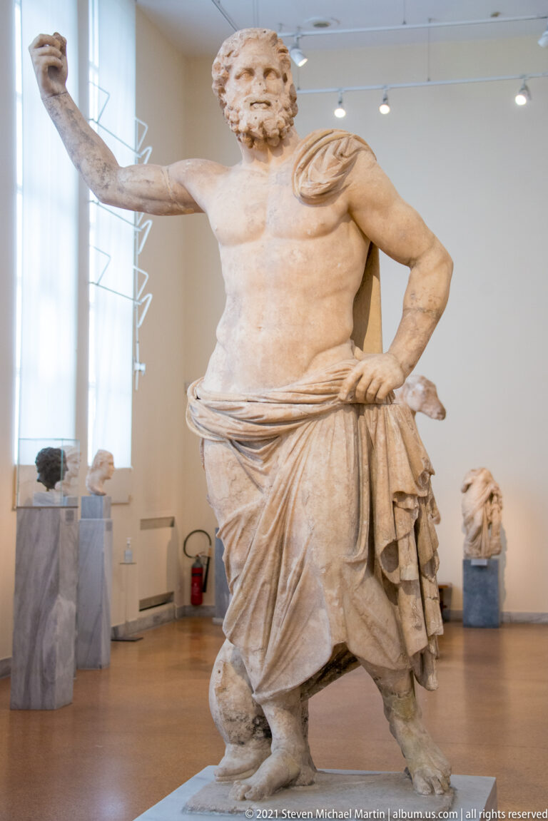 Bearded marble male statue with bare chest, heavy drapery, one hand on the hip, and the opposite arm extended.