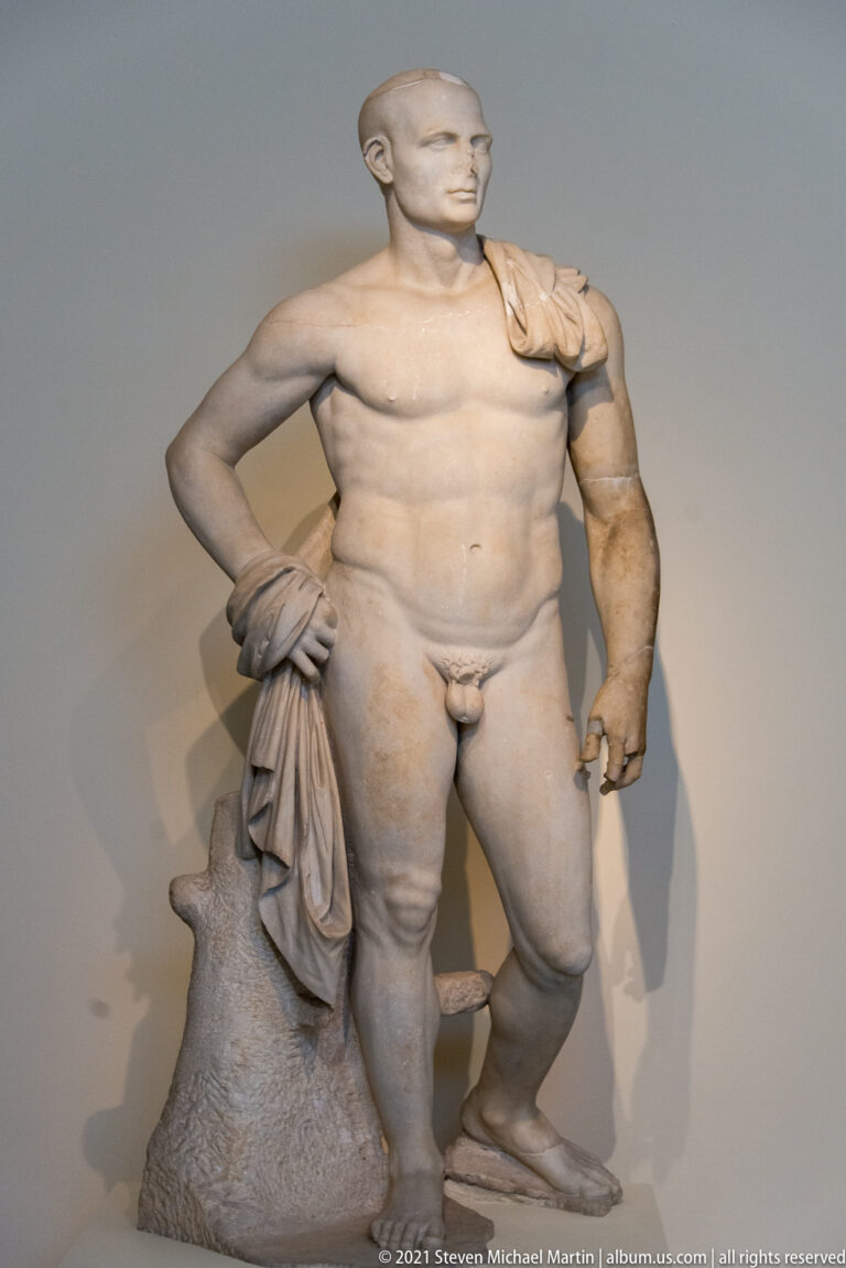 Marble nude male statue with drapery gathered over a support beside one leg and an asymmetrically preserved arm.