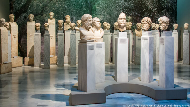 Gallery filled with ancient sculpted heads and busts displayed on tall white pedestals in multiple rows.