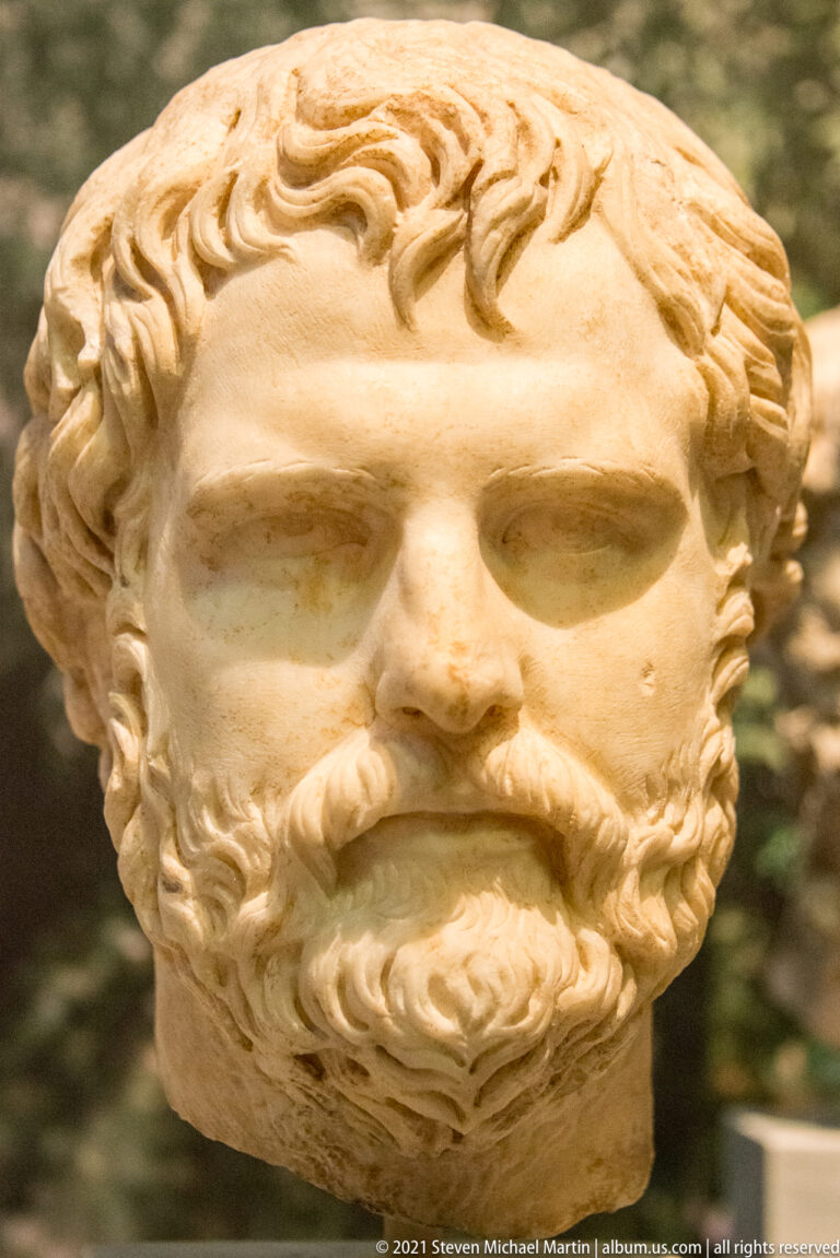 Close view of a marble male head with thick curly beard, swept hair, heavy brow, and deep-set eyes.