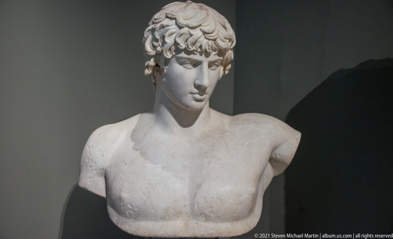 Marble bust-length youthful male figure with curled hair, smooth face, and broken shoulders.