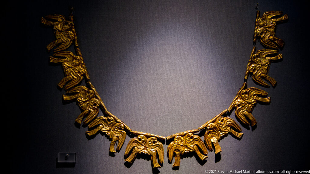 U-shaped gold ornament composed of linked elements and repeated pendant figures or plaques against a dark display wall.