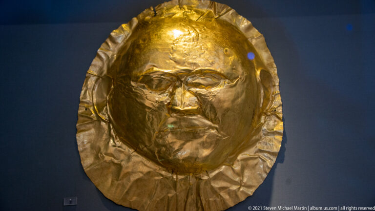Rounded hammered gold funerary mask with modeled male facial features and an irregular circular edge.