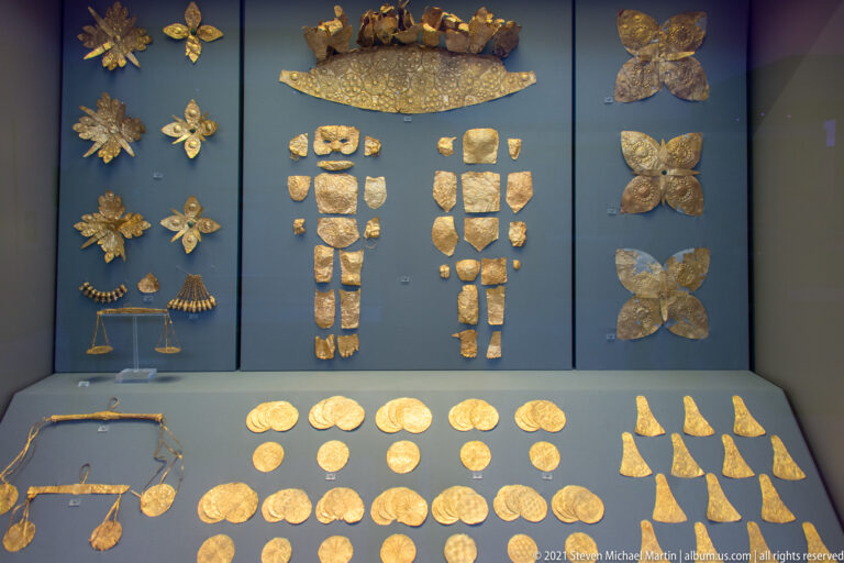 Display case containing grouped Mycenaean gold ornaments, plaques, round disks, pointed pieces, and decorative strips.
