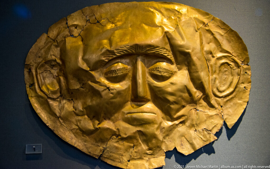 Gold funerary mask with elongated male face, arched brows, large ears, long nose, and irregular damaged edges.