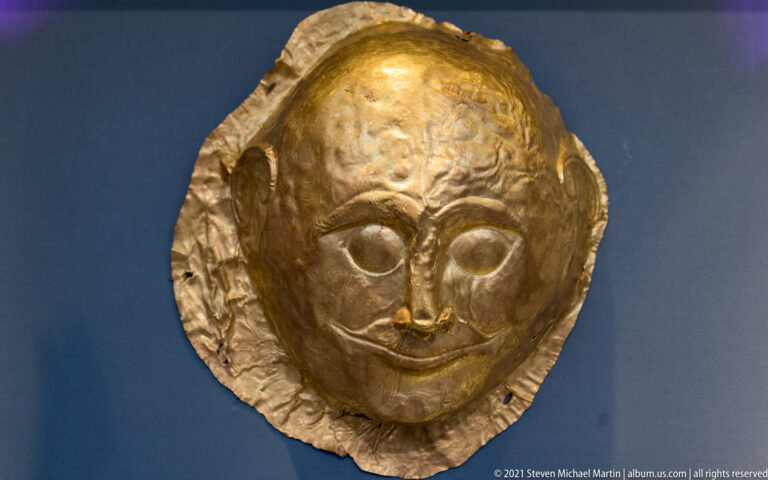 Small rounded gold funerary mask with large almond-shaped eyes, narrow nose, arched brows, and irregular edge.
