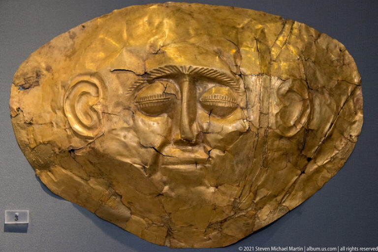 Broad oval gold funerary mask with stylized eyes, large ears, long nose, compressed mouth, and visible cracks.