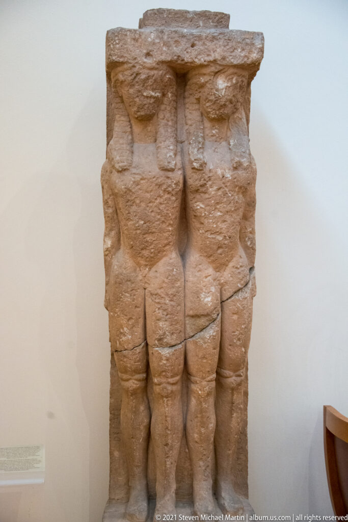 Weathered stone sculpture showing two frontal male figures standing closely side by side.