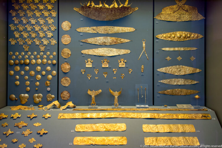 Large museum case filled with grouped Mycenaean gold ornaments, disks, strips, plaques, rosettes, and small appliqués.