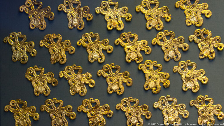 Rows of small gold appliqués with symmetrical curling loop motifs arranged across a dark museum display board.