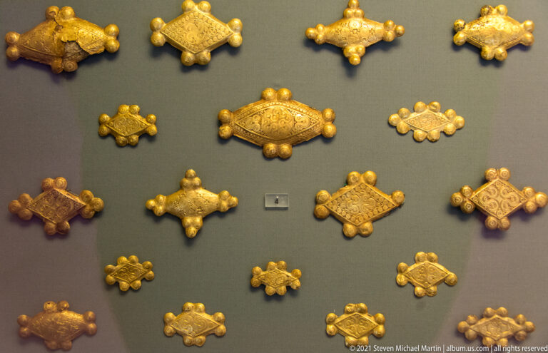 Small gold plaques in diamond and oval shapes with raised bosses and patterned surfaces, arranged on a dark display.