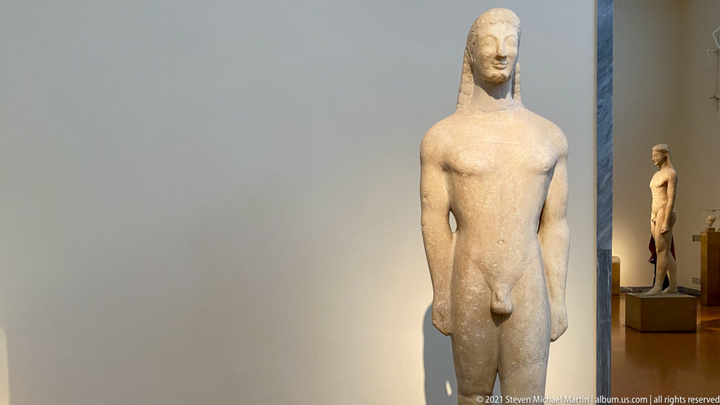 Archaic marble kouros standing nude and frontal with long hair, arms at the sides, and one leg advanced.