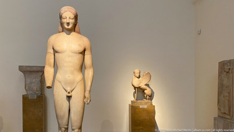 Tall Archaic marble kouros with long segmented hair, frontal nude body, arms at the sides, and one leg advanced.