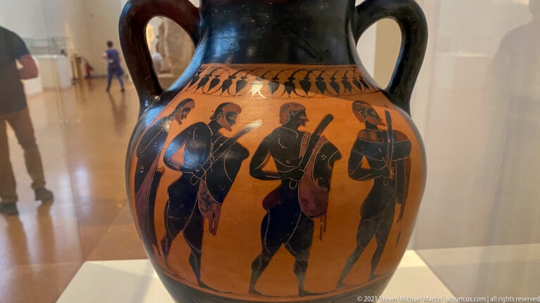 Black-figure amphora detail with a row of standing male figures beneath a decorative leaf band.