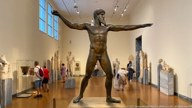 Large bronze nude male statue standing with legs apart and both arms extended horizontally in a museum gallery.