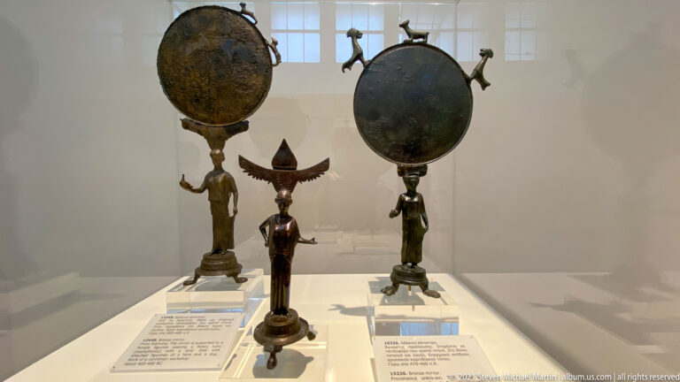 Glass case with tall bronze disk-shaped objects on figural supports and smaller decorative bronze objects between them.