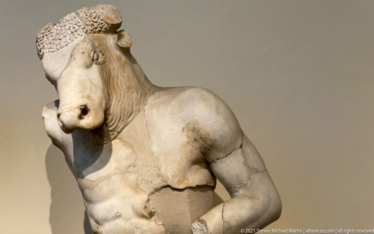 Fragmentary marble bull with broad muzzle, horn, heavy neck folds, and broken torso.