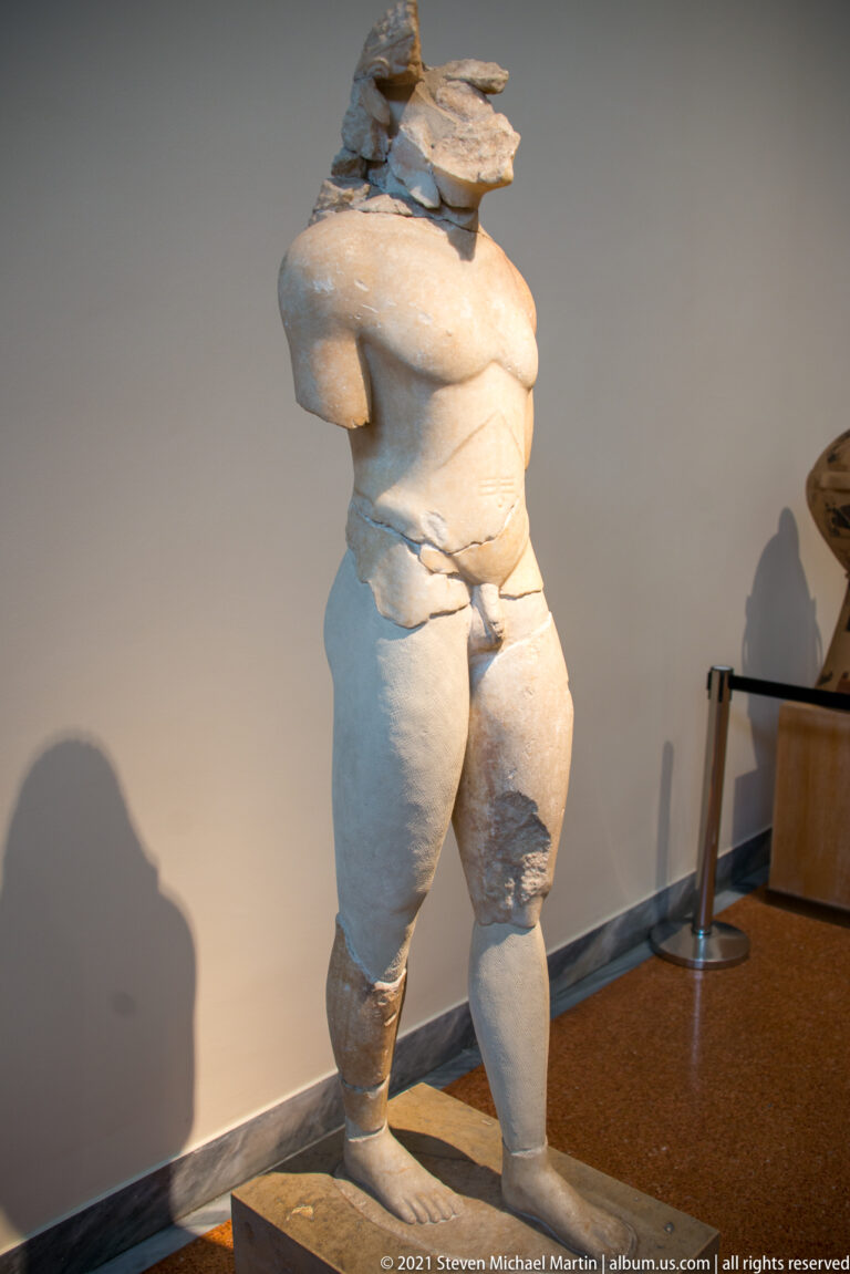 Headless fragmentary kouros with broad chest, modeled abdomen, and damaged legs.