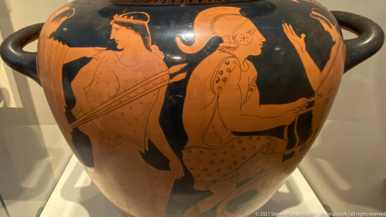 Red-figure handled vessel with armored and draped figures, including helmets, shields, and spears, on a black ground.