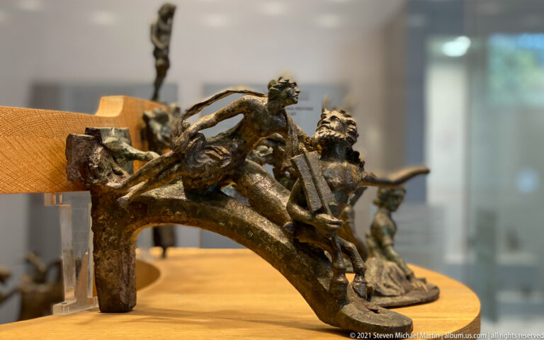 Curved bronze fitting covered with small reclining and standing human figures, displayed on a museum support.