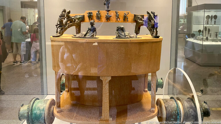 Large cylindrical wooden museum reconstruction with bronze-toned fittings, curved side elements, and small figures across the top.