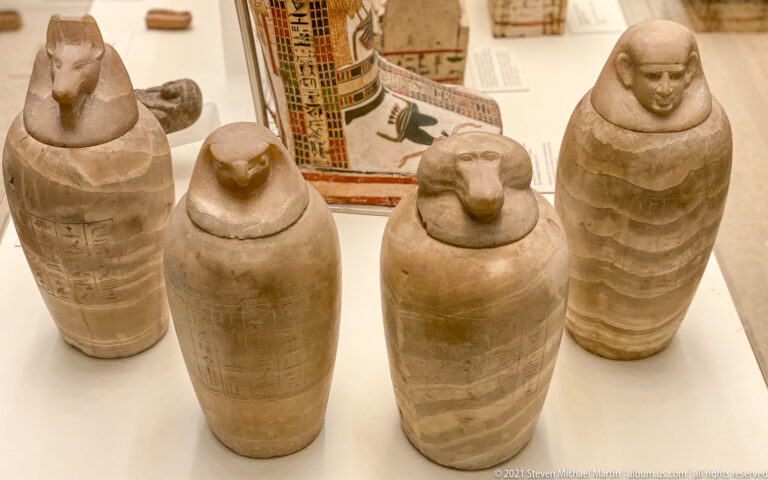 Four pale stone Egyptian canopic jars with human-headed lids displayed together in a museum case.