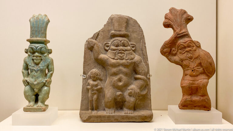Three small protective deity figures in green, stone, and reddish materials, including dwarf-like Bes forms.