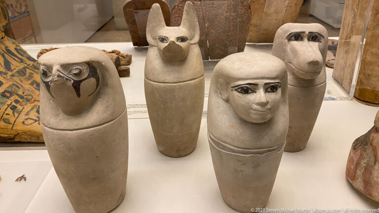 Four Egyptian canopic jars with human, falcon, jackal, and baboon-headed lids grouped in a museum case.